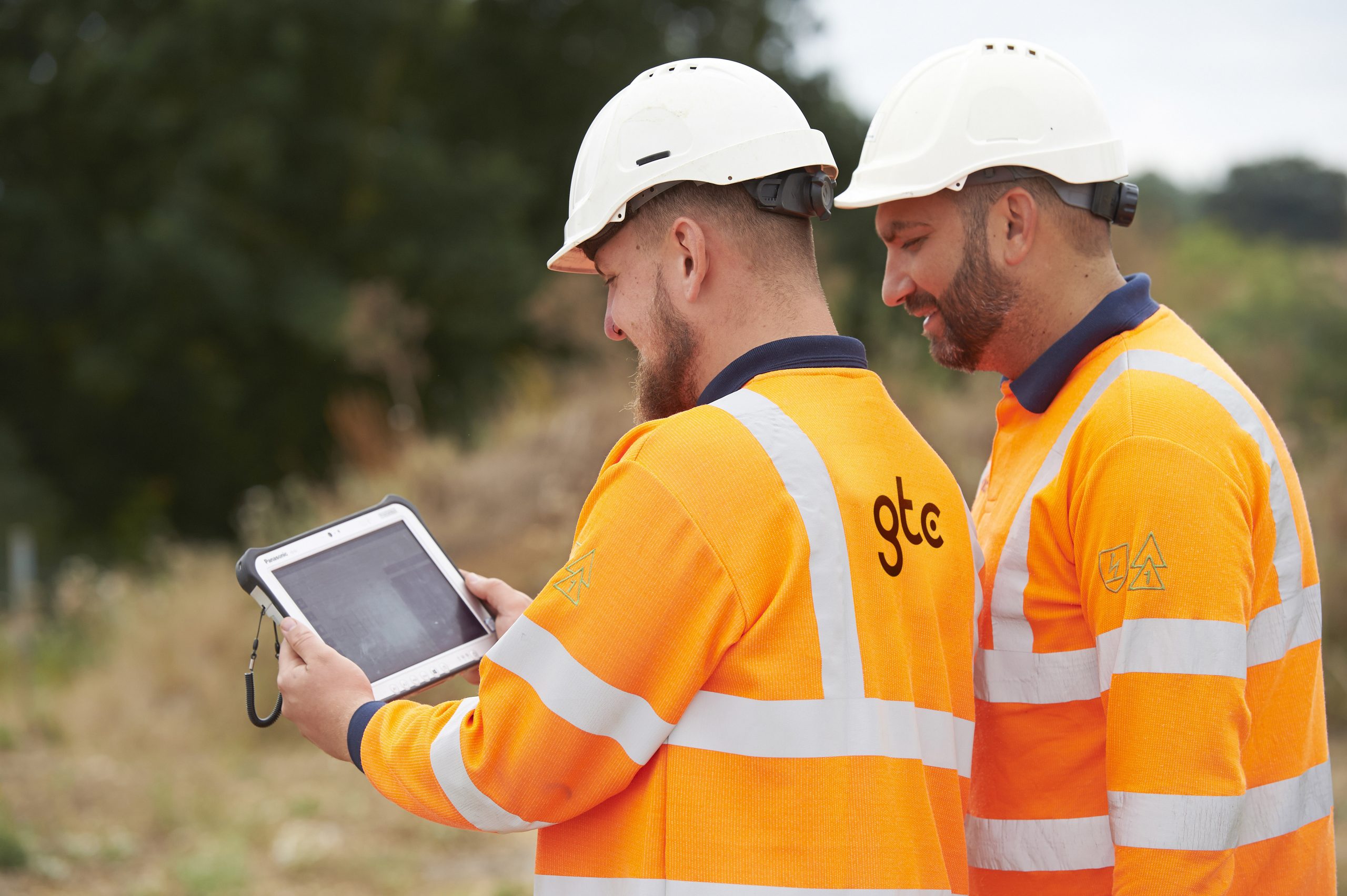 Two men on a construction site, in orange high visibility jackets and hard hats, looking at a tablet together.