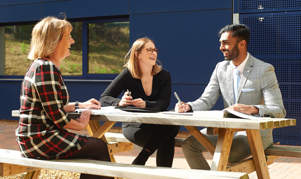 Two women and one man sat on a picnic bench outside an office, they are laughing and smiling at each other.