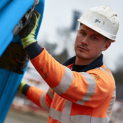 A construction worker in an orange high visibility jacket and hard hat, rolling a large circle of blue pipe along the ground.